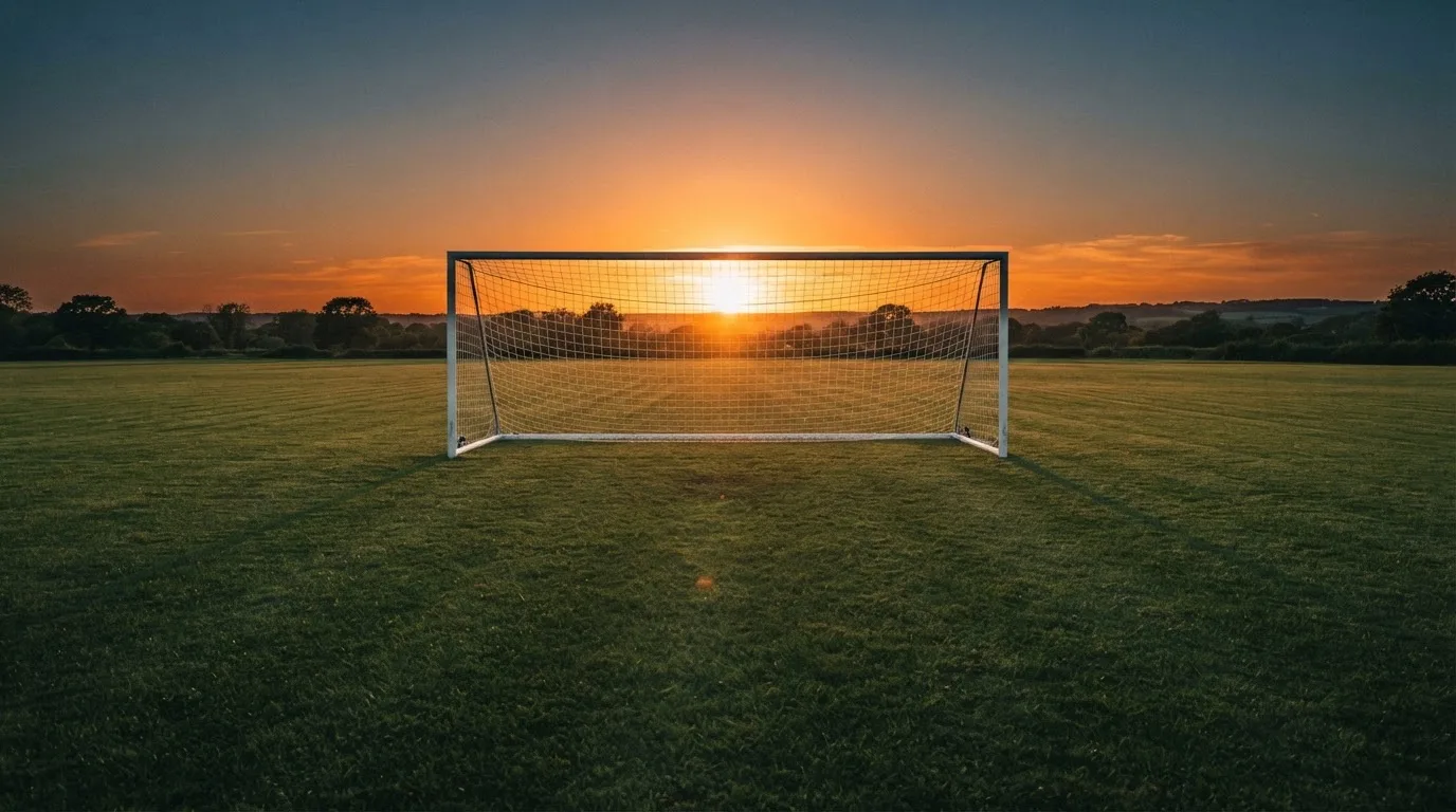 Ein Fußballtor mit Netz bei Sonnenuntergang auf einem leeren Platz, die Schatten der Torpfosten liegen lang auf dem Gras.