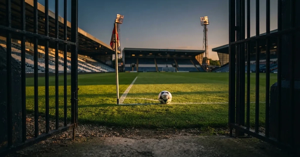 Blick durch ein Stadiontor auf einen grünen Rasen im Abendlicht, ein Ball ruht an der Eckfahne, die Tribünen sind leer.