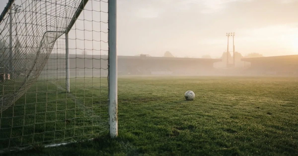 Ein Fußballtor mit gespanntem Netz steht auf einem leeren Rasenplatz, davor ein einzelner Ball, Morgendunst liegt über dem Feld.