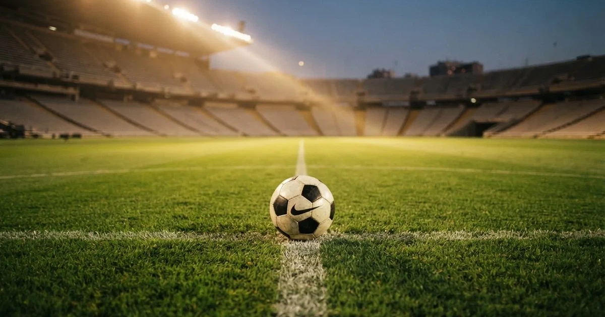 Ein Fußball liegt auf dem Rasen eines leeren Stadions bei Flutlicht, im Hintergrund unscharfe Tribünen ohne erkennbare Beschriftung.