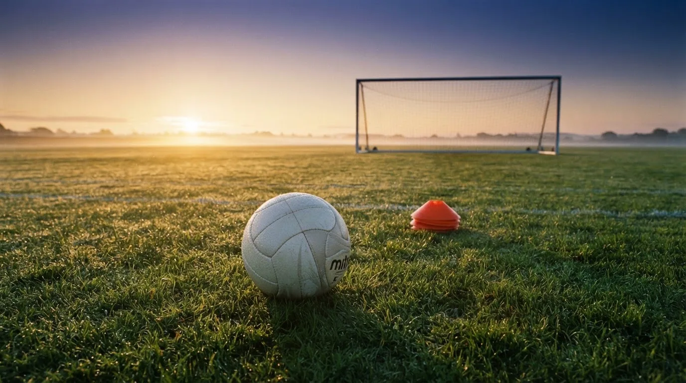 Ein Fußball liegt auf einem Rasenplatz bei Sonnenaufgang, daneben ein kleiner Stapel Trainingshütchen.
