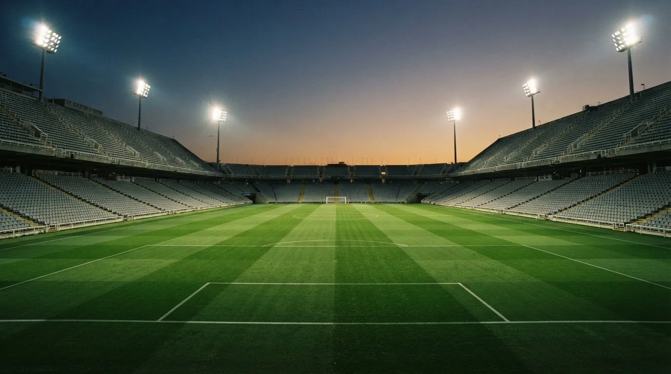 Eine leere Stadiontribüne unter Flutlicht aus der Ferne betrachtet, die Rasenfläche leuchtet grün in der Dämmerung.