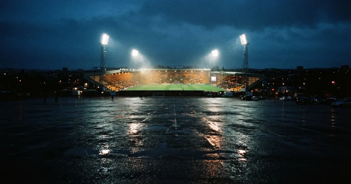Ein Stadion unter Flutlicht aus großer Entfernung gesehen, davor ein regennasser Parkplatz in der Dämmerung.