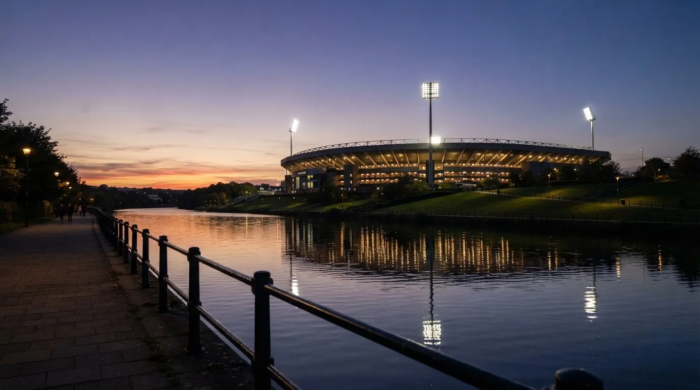 Ein Stadion unter Flutlicht aus der Ferne gesehen, im Vordergrund ein ruhiger Fluss und ein Fußweg.