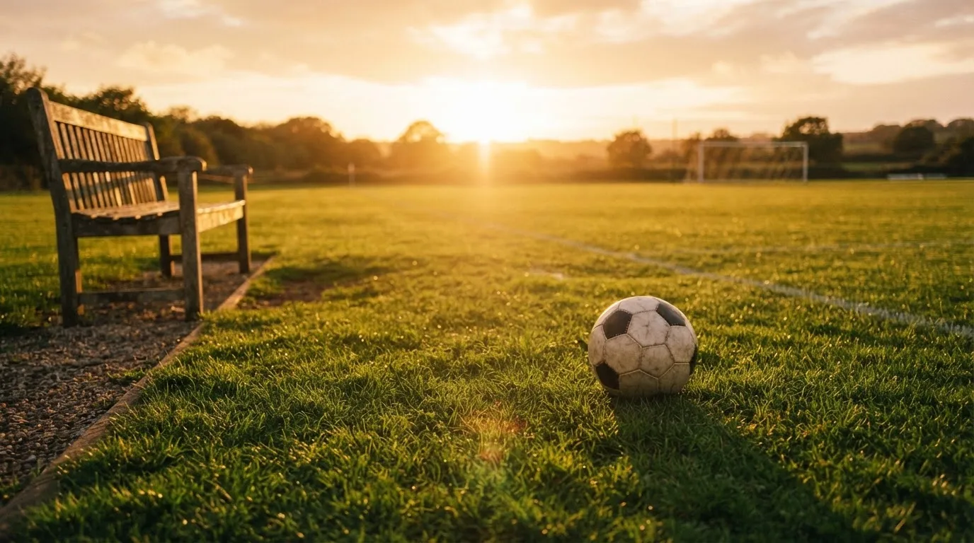 Ein Fußball auf einem Rasenplatz im Gegenlicht der tiefstehenden Sonne, am Rand eine leere Ersatzbank.