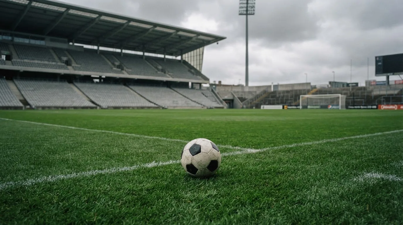 Ein Fußball auf einem Rasen, im Hintergrund ein leeres Stadion bei bewölktem Himmel.