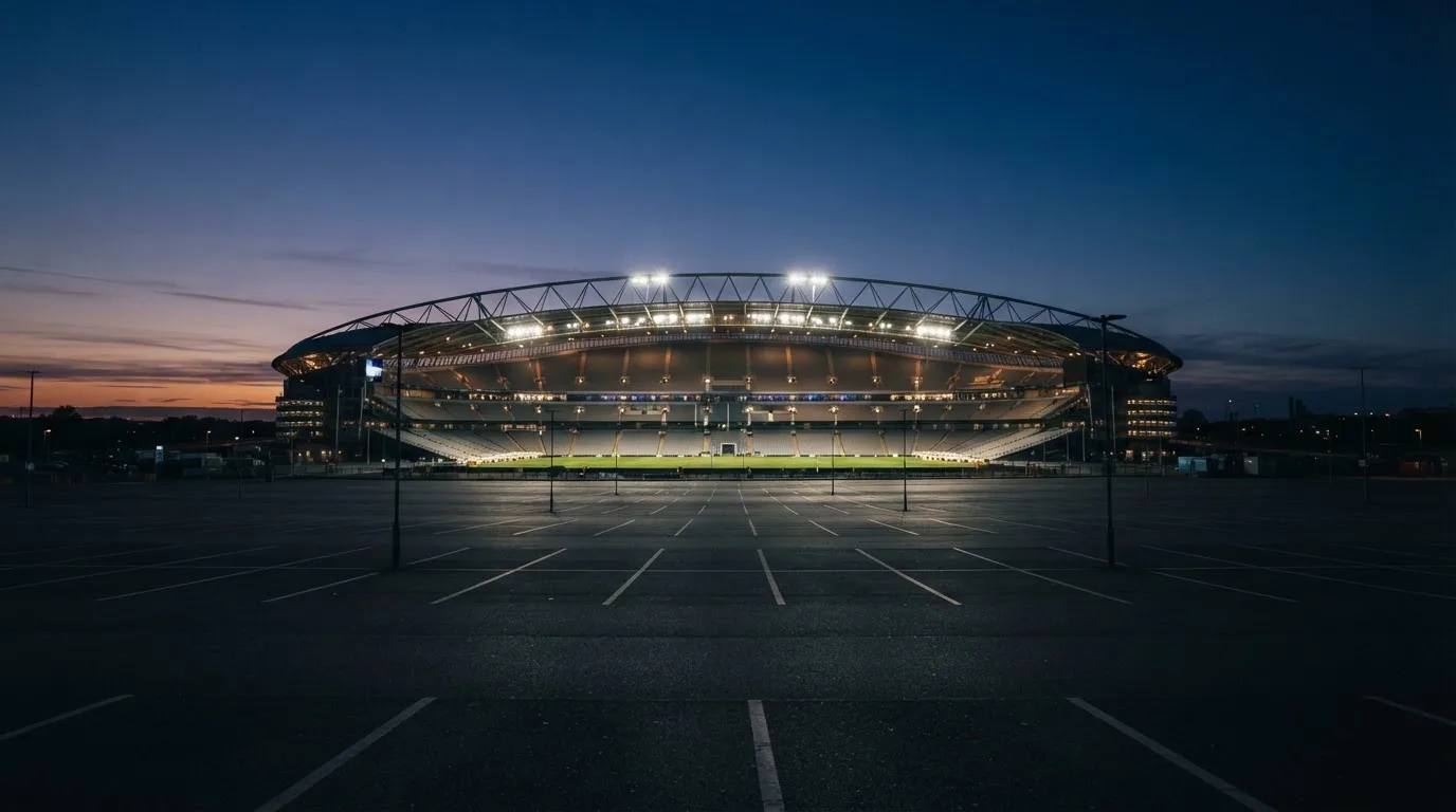 Ein Fußballstadion unter Flutlicht, aus großer Entfernung bei Einbruch der Dunkelheit gesehen, davor ein leerer Parkplatz.