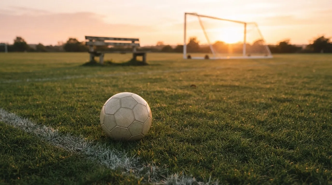 Ein Fußball liegt im Gras am Spielfeldrand, dahinter eine leere Bank und ein Tor im Gegenlicht.