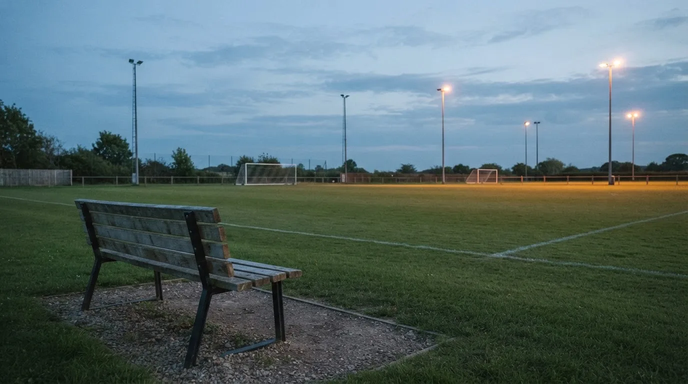 Eine leere Parkbank am Rand eines Fußballfelds in der Abenddämmerung, im Hintergrund ragen dunkle Flutlichtmasten in den Himmel.
