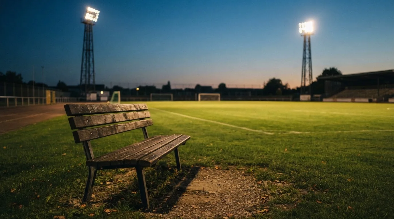 Eine leere Parkbank am Rand eines Fußballfelds in der Abenddämmerung, im Hintergrund ragen dunkle Flutlichtmasten in den Himmel.