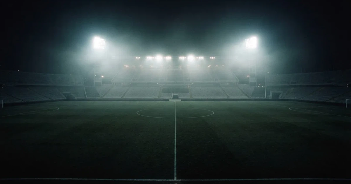 Ein Stadion unter Flutlicht, aus großer Entfernung über eine dunkle Wiese fotografiert, am Himmel zieht Nebel auf.