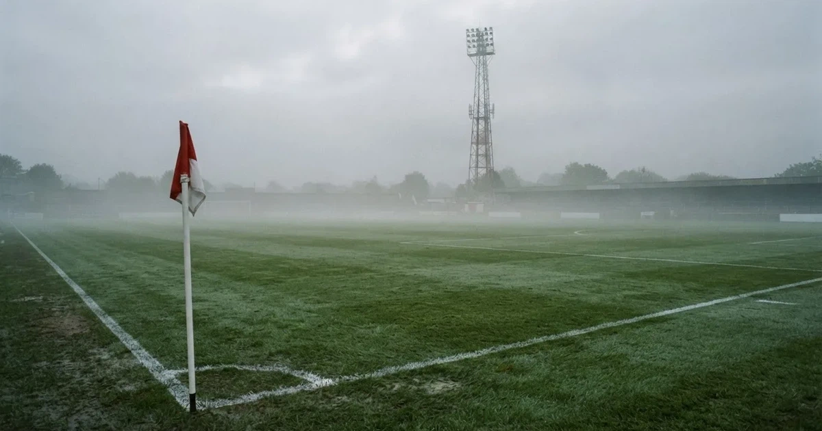 Eine Eckfahne steht im Nebel am Rand eines leeren Rasenplatzes in der Morgendämmerung, dahinter ein Flutlichtmast.