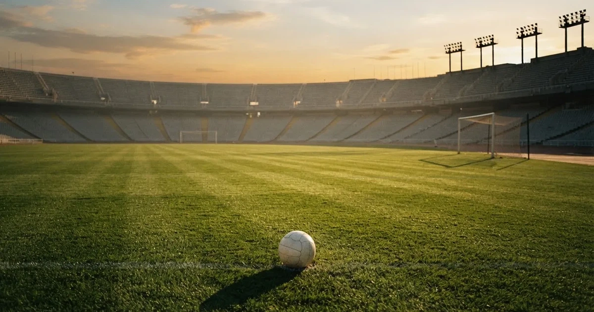 Ein Fußballplatz mit frisch gemähtem Rasen im Abendlicht, ein Ball liegt im Strafraum, im Hintergrund unscharf eine leere Tribüne.