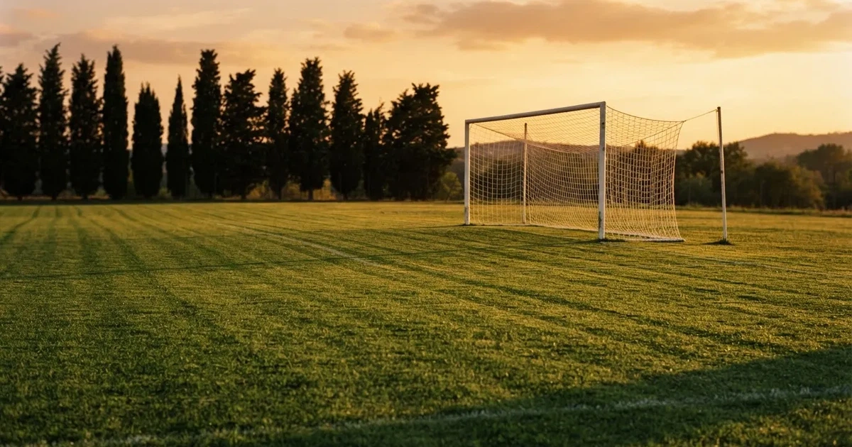 Ein Fußballtor mit Netz auf einem leeren Platz im Abendlicht, der Rasen frisch gemäht, im Hintergrund Zypressen.