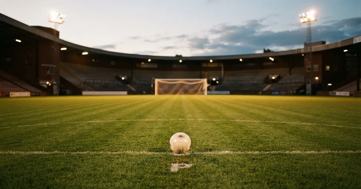 Ein Fußballplatz mit frisch gemähtem Rasen im Abendlicht, ein Ball liegt im Strafraum, im Hintergrund unscharf eine leere Tribüne.