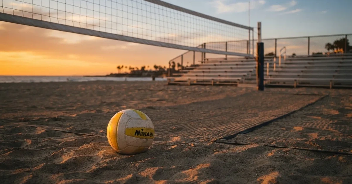 Ein Volleyball liegt auf dem Sand eines Beachvolleyball-Feldes neben dem gespannten Netz, im Hintergrund eine leere Tribüne bei Abendlicht.