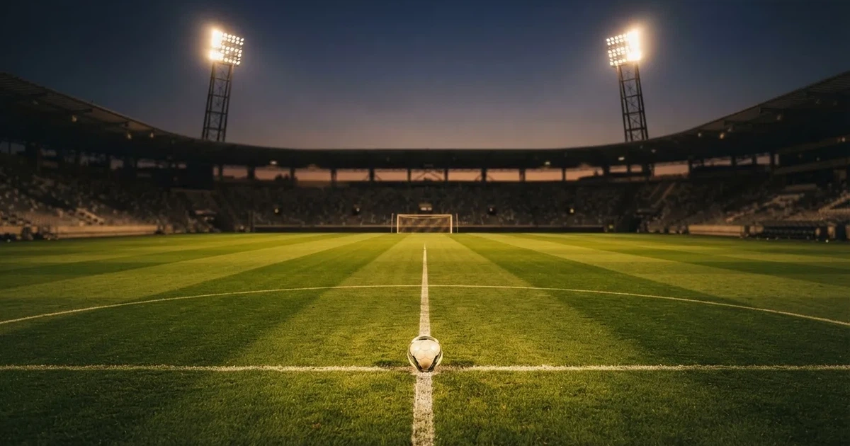 Ein Rasenplatz in der Dämmerung mit eingeschalteten Flutlichtmasten, ein Ball liegt am Anstoßpunkt, die Tribünen im Hintergrund sind unscharf.
