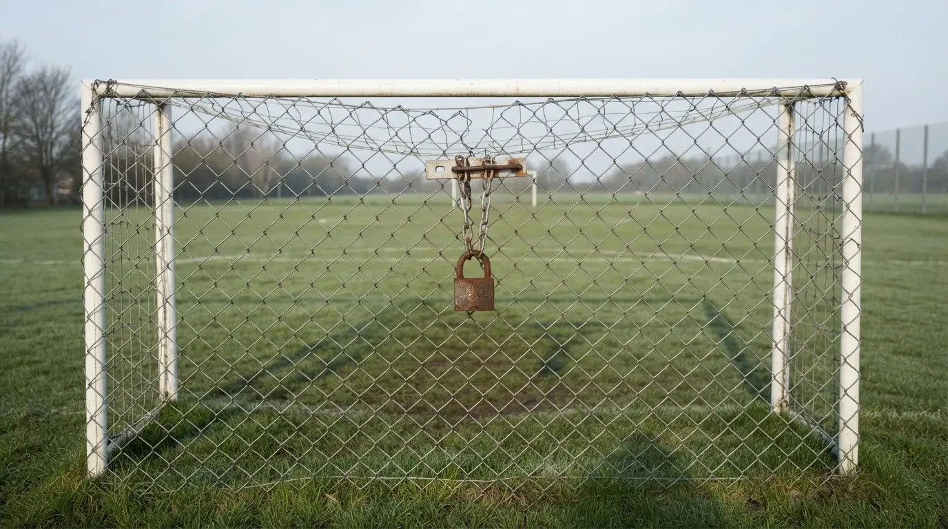 Ein geschlossenes Tor an einem Zaun mit Vorhängeschloss im Morgenlicht, dahinter ein einfacher Sportplatz.