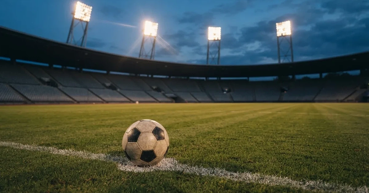 Ein Fußball liegt auf dem Anstoßpunkt eines leeren Rasenplatzes, im Hintergrund eine Tribüne und Flutlichtmasten in der Abenddämmerung.