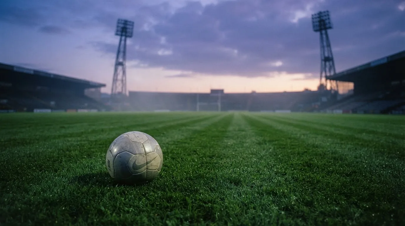 Ein Fußball liegt im Gras eines leeren Platzes, im Hintergrund stehen Flutlichtmasten in der Dämmerung.