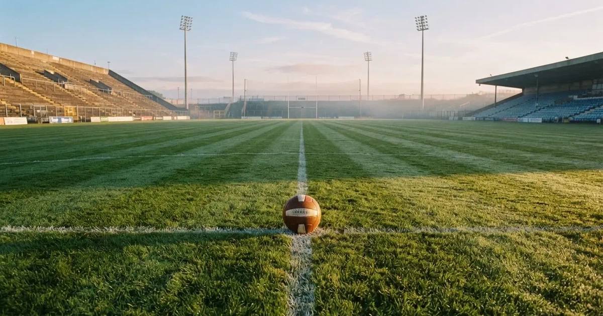 Ein Fußball liegt auf dem Anstoßpunkt eines Rasenplatzes, dahinter leere Tribünen im Licht des frühen Morgens.