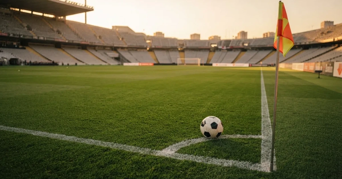 Ein Fußball liegt am Viertelkreis an der Eckfahne eines Rasenplatzes, im Hintergrund leere Tribünen in warmem Abendlicht.