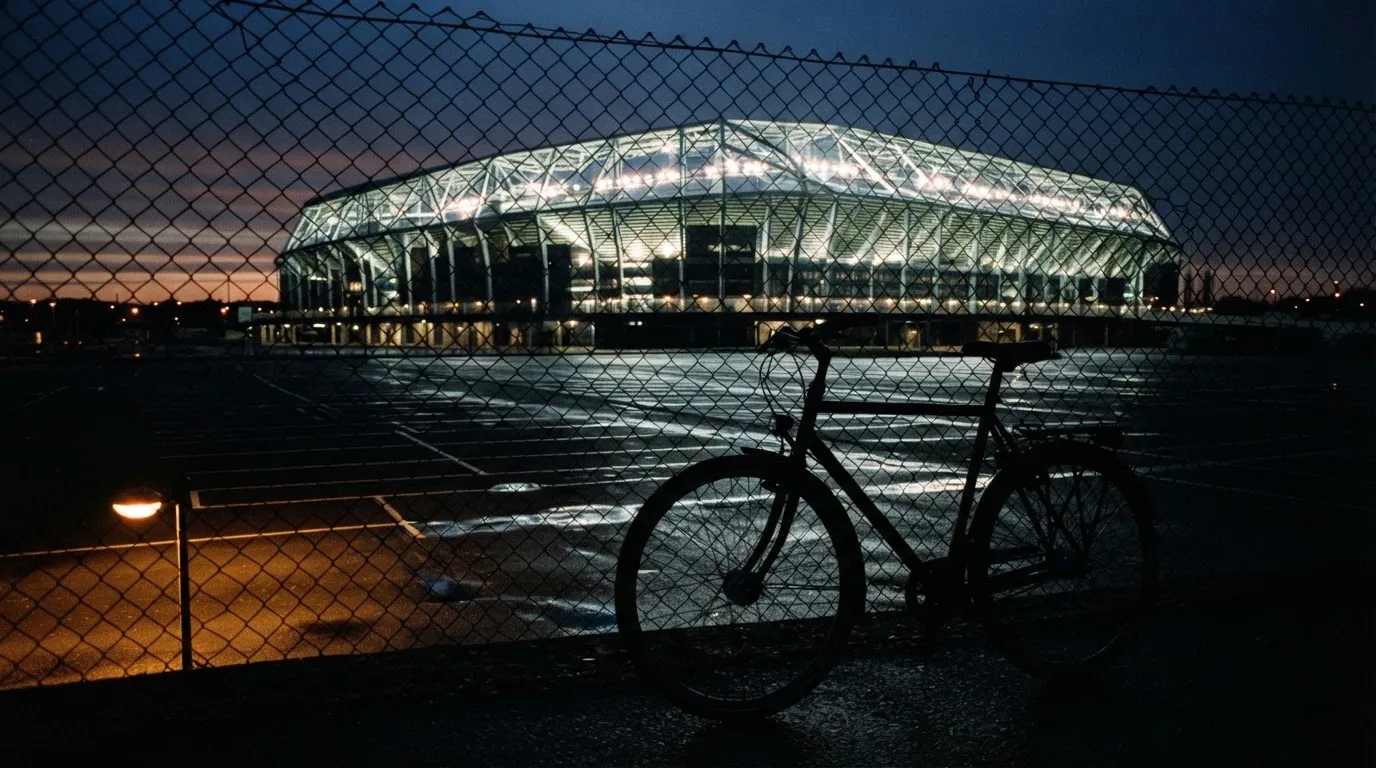 Ein Stadion unter Flutlicht aus der Ferne gesehen, davor ein dunkler Parkplatz und ein einzelnes Fahrrad an einem Zaun.