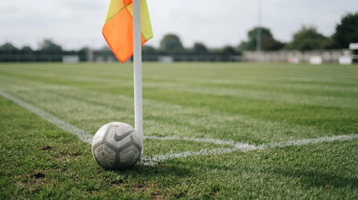 Ein Fußball liegt neben einer Eckfahne auf kurz gemähtem Rasen unter bewölktem Himmel.